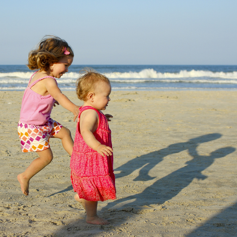 Shove: Big sister leaping into the air to push little sister into the sand. Beach with waves in background. By Calm Cradle Photo & Design